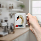 A hand holds the Nordic Wholesale Mug: Denmark National Animal Mute Swan Ceramic Mug (11 oz)—a crisp white piece featuring an elegant mute swan, the Danish flag, and the inscription "DENMARK Knopsvane | Mute Swan"—in a bright, modern kitchen.