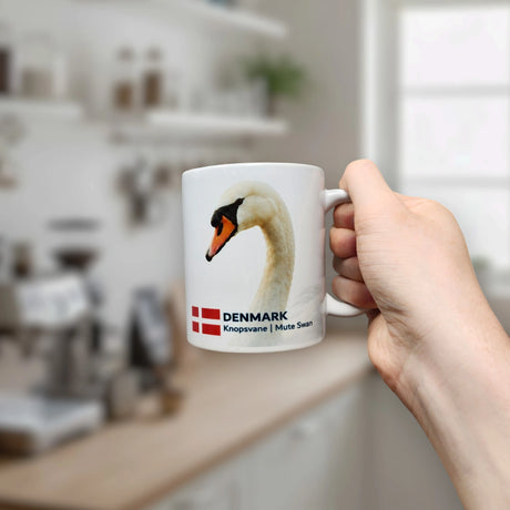 A hand holds the Nordic Wholesale Mug: Denmark National Animal Mute Swan Ceramic Mug (11 oz)—a crisp white piece featuring an elegant mute swan, the Danish flag, and the inscription "DENMARK Knopsvane | Mute Swan"—in a bright, modern kitchen.