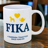 A white ceramic Mug: Fika — A Swedish Tradition Since Forever (11 oz) by Nordic Wholesale rests on a wooden table, featuring the phrase "FIKA: A Swedish tradition since forever" and icons of a yellow horse, heart, and teapot above the text.
