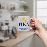 A hand holds the Nordic Wholesale Mug: Fika — A Swedish Tradition Since Forever Ceramic Mug (11 oz), featuring blue and yellow designs, in a blurred kitchen setting.