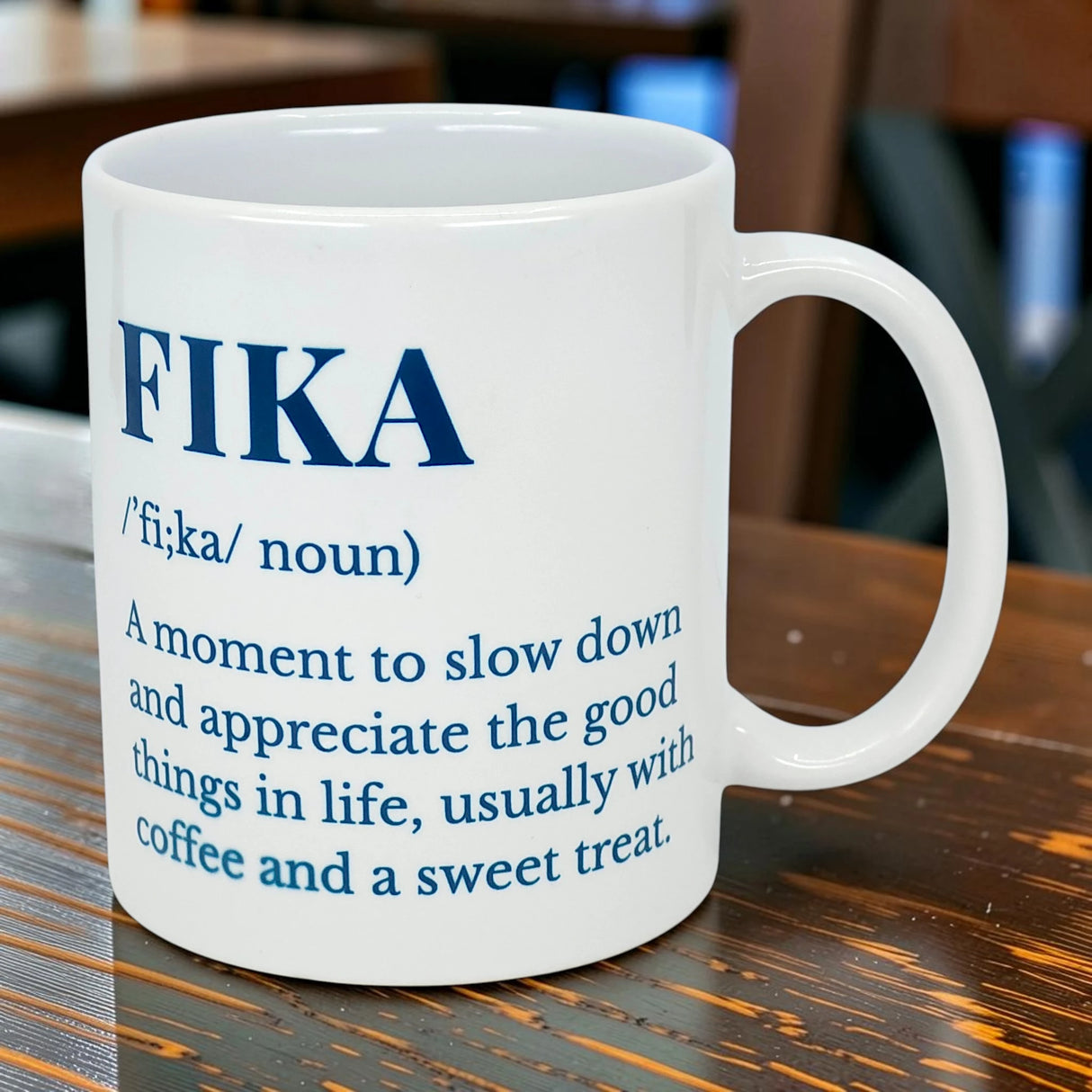 The Swedish Gift Store's White Ceramic Fika Definition Mug (11 oz) sits on a wooden table, featuring the meaning of Fika—a time to slow down and enjoy life, often with coffee and a sweet treat.