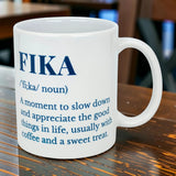 The Swedish Gift Store's White Ceramic Fika Definition Mug (11 oz) sits on a wooden table, featuring the meaning of Fika—a time to slow down and enjoy life, often with coffee and a sweet treat.