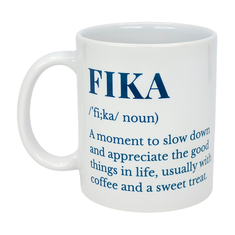 The Nordic Wholesale White Ceramic Fika Definition Mug (11 oz) features blue text defining FIKA as a moment to slow down and enjoy coffee and treats.