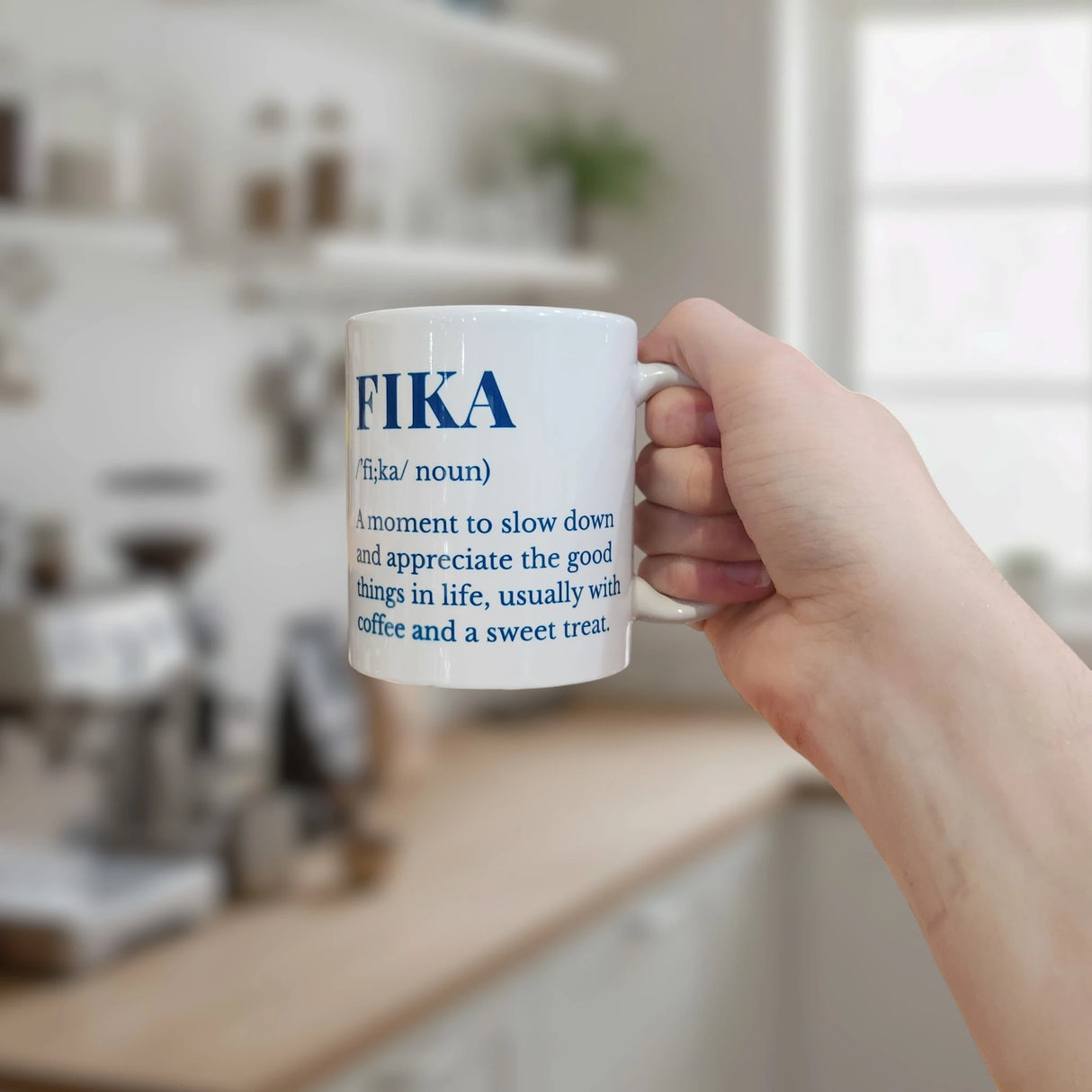A hand holds the Swedish Gift Store's White Ceramic Fika Definition Mug (11 oz), featuring blue text about slowing down to enjoy life's good moments—often with coffee and a sweet—in a bright, modern kitchen.