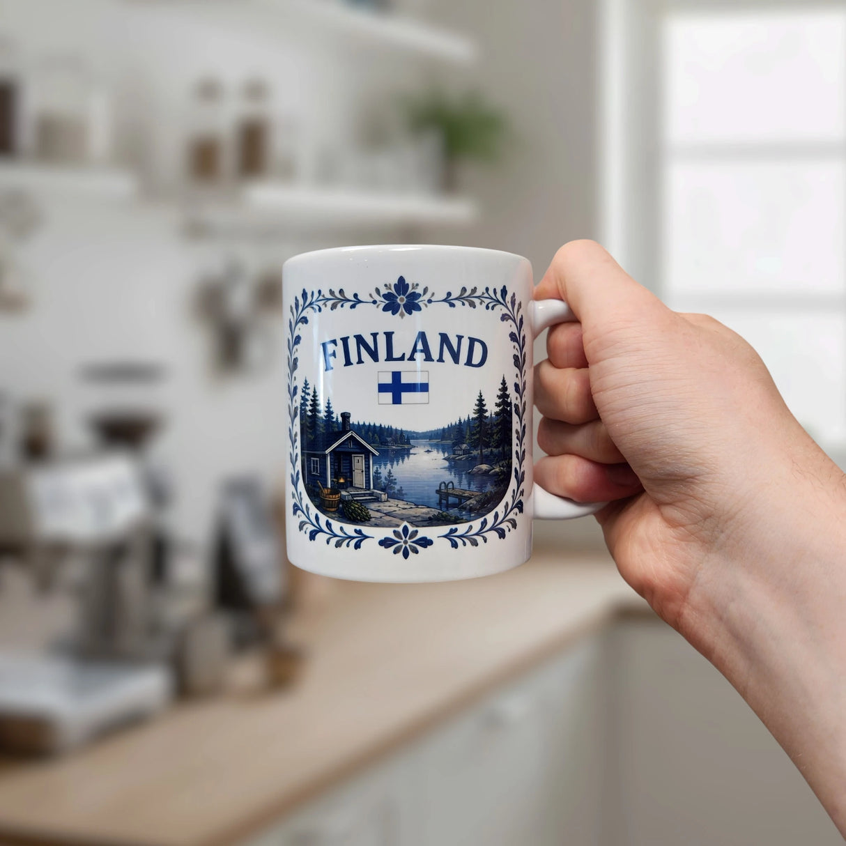 A hand holds the "Finland Lakeside Cabin Ceramic Mug" (11 oz capacity) by Nordic Wholesale, featuring a Finnish flag, the word "Finland," blue floral designs, and an illustration of a Nordic lakeside cabin; a blurred kitchen forms the background.
