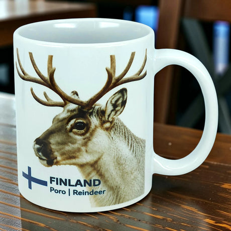A white ceramic mug from Nordic Wholesale, featuring Finland’s national animal—the reindeer—along with a blue Finnish flag and the label "FINLAND Poro | Reindeer," sits on a wooden table. This mug has an 11 oz capacity.