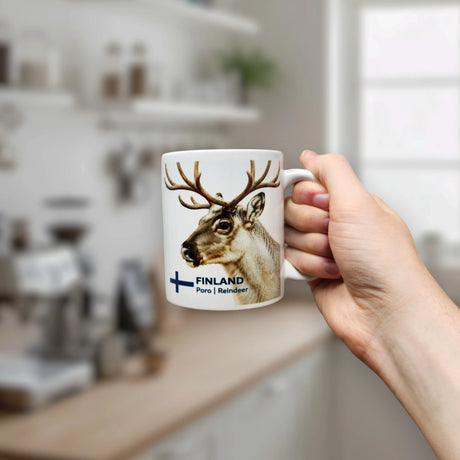 A hand holds the Nordic Wholesale Mug: Finland National Animal Reindeer Ceramic Mug (11 oz Capacity), which features an image of a reindeer and the words "FINLAND" and "Poro | Reindeer." In the background, a softly blurred kitchen with shelves and a window accentuates the cozy Nordic charm of this distinctive ceramic mug.