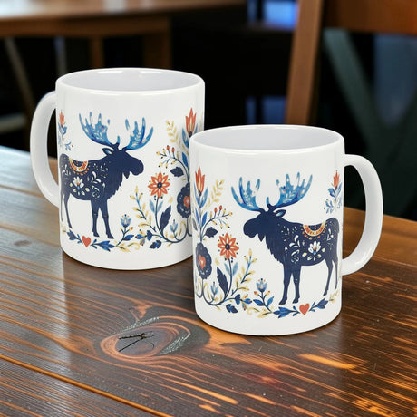 Two Nordic Wholesale Folk Art Moose Ceramic Mugs (11 oz) with white glaze feature blue moose and vibrant Scandinavian floral patterns in blue, orange, and yellow, inspired by Nordic folk art. Perfect ceramic coffee cups for your home.