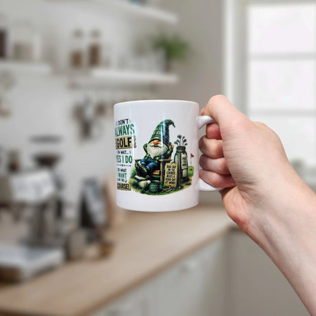 A hand holds the Nordic Wholesale Mug: Funny Tomte Gnome Golf Ceramic Mug (11 oz), featuring a cartoon golf gnome and the quote, "I don’t always play golf… oh wait, yes I do," with a blurred modern kitchen in the background.