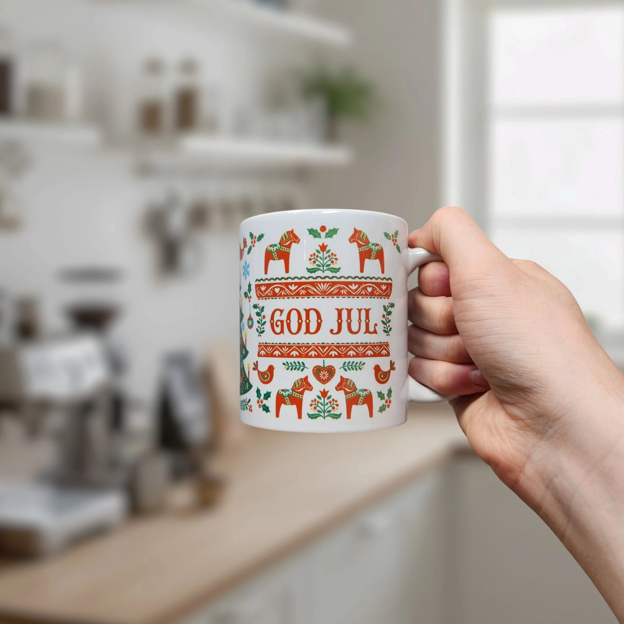 A hand holds the Nordic Wholesale Mug: God Jul Dala Horse Christmas Ceramic Mug (11 oz Capacity), decorated with Scandinavian-style patterns, including red Dala horses, green leaves, and the words "GOD JUL." A blurred kitchen can be seen in the background.