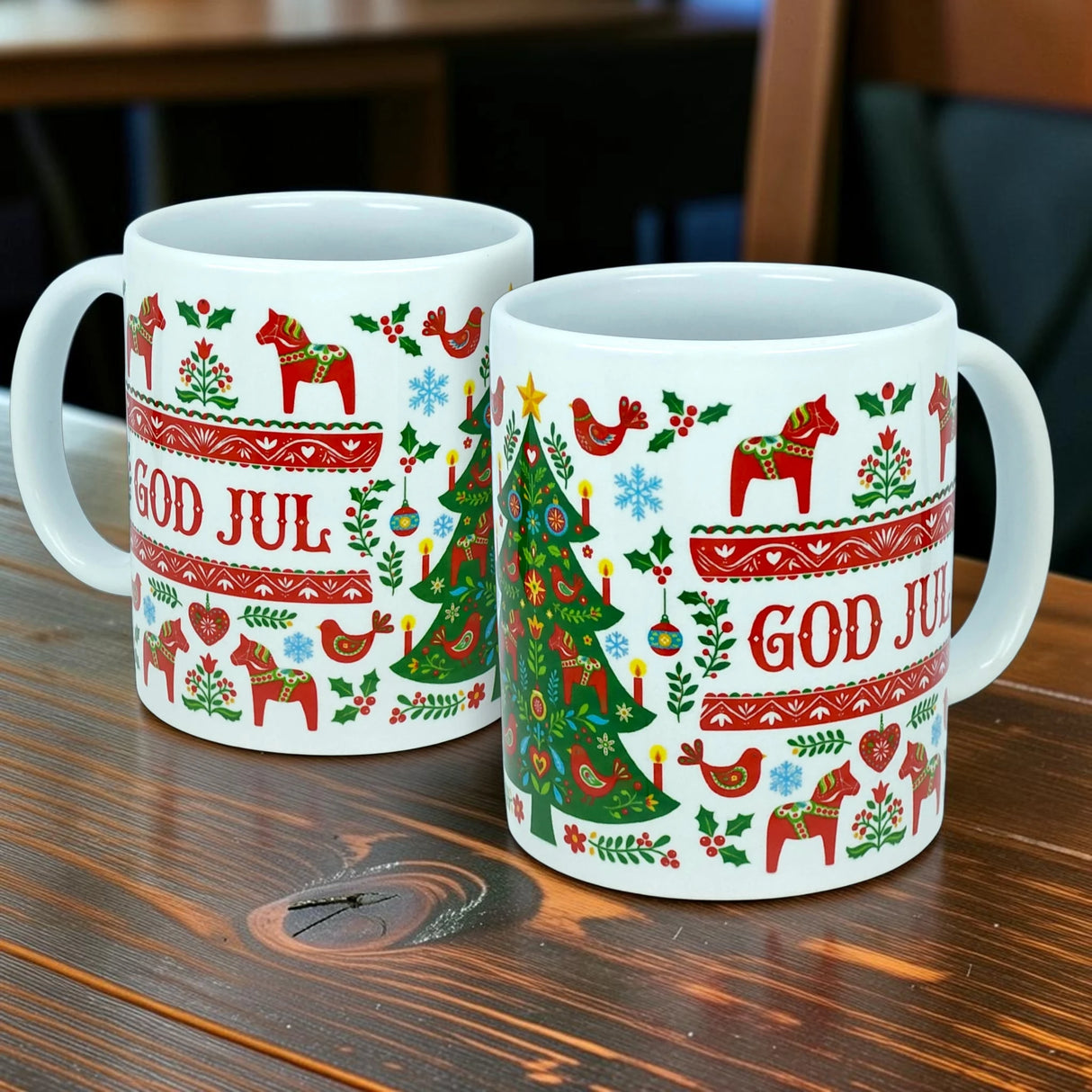 Two white Mug: God Jul Dala Horse Christmas Ceramic Mugs (11 oz capacity) from Nordic Wholesale, featuring red text and charming Scandinavian holiday designs—including horses, birds, snowflakes, and a Christmas tree—sit on a wooden table. These festive mugs bring the warmth and beauty of Nordic tradition to your holiday gatherings.