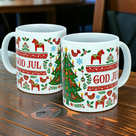 Two white Mug: God Jul Dala Horse Christmas Ceramic Mugs (11 oz capacity) from Nordic Wholesale, featuring red text and charming Scandinavian holiday designs—including horses, birds, snowflakes, and a Christmas tree—sit on a wooden table. These festive mugs bring the warmth and beauty of Nordic tradition to your holiday gatherings.