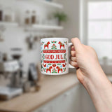 A hand holds the Nordic Wholesale Mug: God Jul Dala Horse Christmas Ceramic Mug (11 oz Capacity), a white Scandinavian-style mug decorated with red and green Nordic patterns and the words "God Jul," in a kitchen setting with blurry shelves and a window in the background.