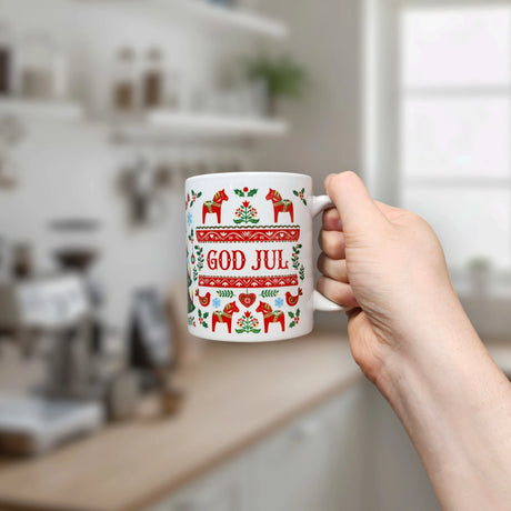A hand holds the Nordic Wholesale Mug: God Jul Dala Horse Christmas Ceramic Mug (11 oz Capacity), a white Scandinavian-style mug decorated with red and green Nordic patterns and the words "God Jul," in a kitchen setting with blurry shelves and a window in the background.