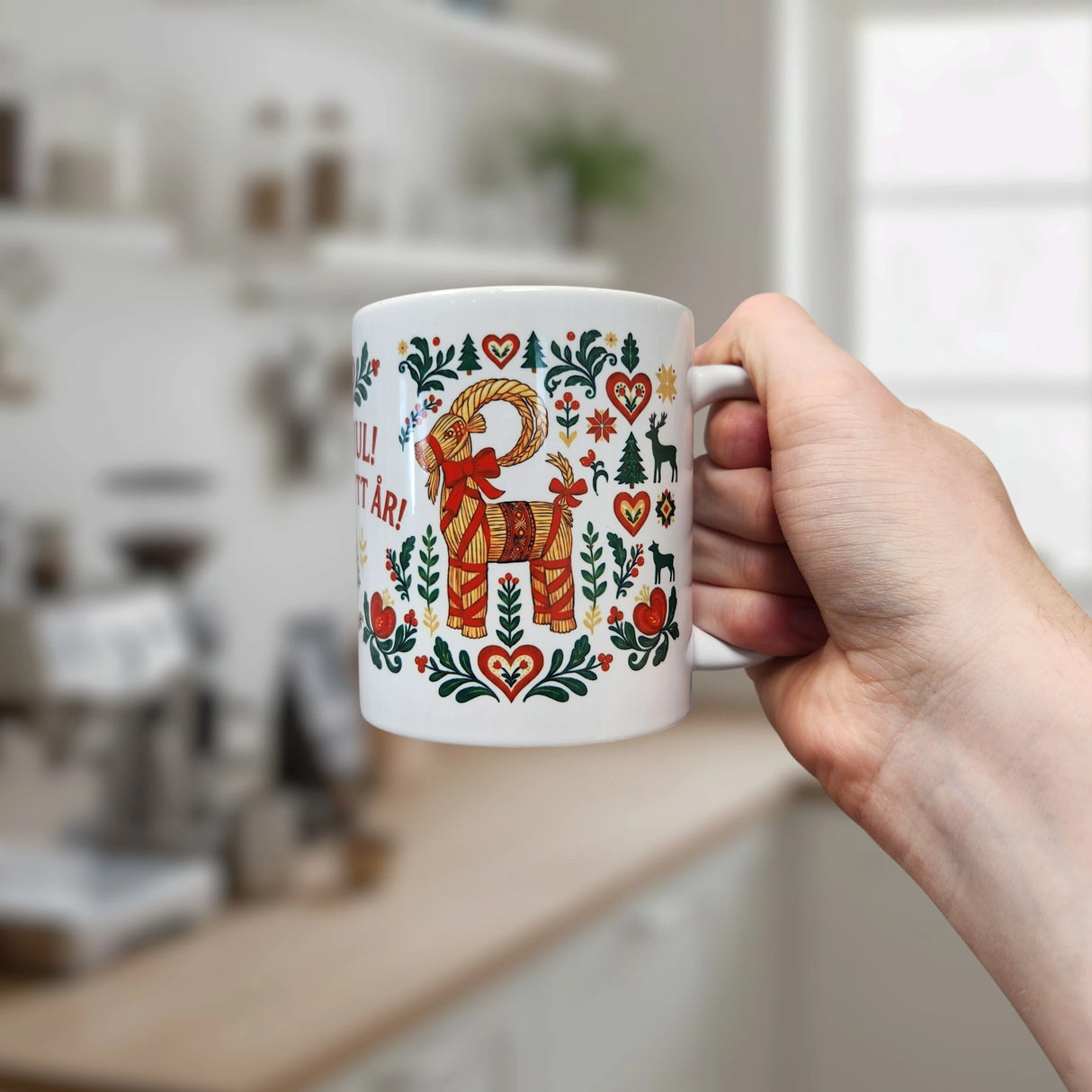 A hand holds a Nordic Wholesale Mug: God Jul Gott Nytt År Yule Goat Christmas Ceramic Mug (11 oz capacity), featuring a red and gold yule goat surrounded by heart and floral patterns, with a blurred kitchen in the background.