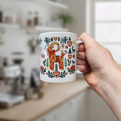 A hand holds a Nordic Wholesale Mug: God Jul Gott Nytt År Yule Goat Christmas Ceramic Mug (11 oz capacity), featuring a red and gold yule goat surrounded by heart and floral patterns, with a blurred kitchen in the background.