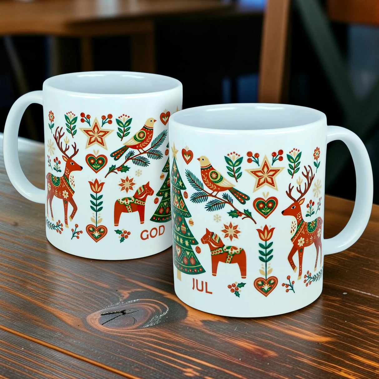 Two God Jul Nordic Folk Art Ceramic Mugs (11 oz) from Nordic Wholesale, featuring white Scandinavian design with colorful folk art motifs of reindeer, birds, hearts, and stars. The mugs display the festive "GOD JUL" greeting—meaning Merry Christmas—and are pictured resting on a wooden table.