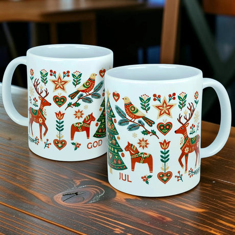 Two God Jul Nordic Folk Art Ceramic Mugs (11 oz) from Nordic Wholesale, featuring white Scandinavian design with colorful folk art motifs of reindeer, birds, hearts, and stars. The mugs display the festive "GOD JUL" greeting—meaning Merry Christmas—and are pictured resting on a wooden table.