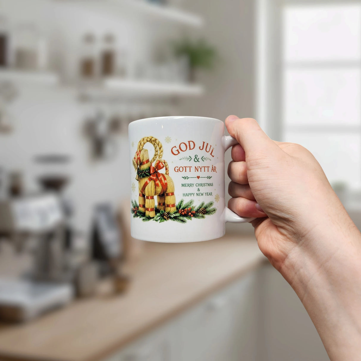 A hand holds the Nordic Wholesale "God Jul Yule Goat Ceramic Mug" (11 oz capacity), a white mug featuring festive text—God Jul & Gott Nytt År, Merry Christmas, Happy New Year—and a classic Scandinavian Yule Goat design. This charming mug brings Swedish Julbock tradition to your holiday celebrations, standing out against the blurred kitchen background.