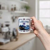 A hand holds the Mug: Iceland Church Mountain Ceramic Mug (11 oz Capacity) by Nordic Wholesale, which features the word "Iceland," an Icelandic flag, and a scenic Nordic landscape. In the background, a softly lit kitchen with shelves and jars is blurred as natural light streams through a window.
