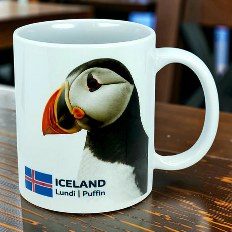 A white ceramic mug by Nordic Wholesale, the "Mug: Iceland National Animal Puffin Ceramic Coffee Mug (11 oz Capacity)," features a charming puffin illustration, the Icelandic flag, and the words "ICELAND Lundi | Puffin." Perfect for Nordic wildlife lovers or anyone who wants to celebrate Iceland's iconic national animal with every sip.
