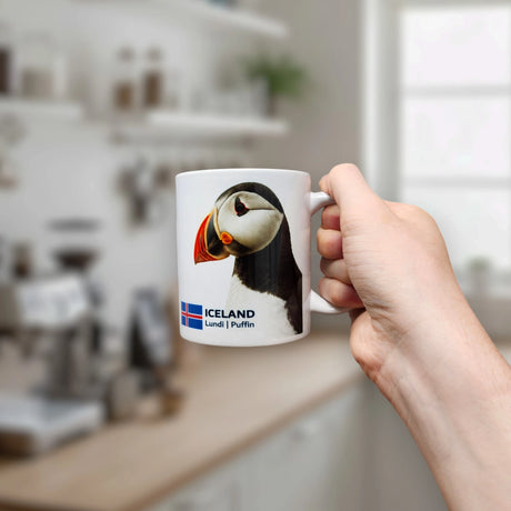 In a bright, modern kitchen setting, a hand holds the Nordic Wholesale Mug: Iceland National Animal Puffin Ceramic Coffee Mug (11 oz Capacity), featuring a charming puffin illustration, the Icelandic flag, and the words ICELAND Lund | Puffin—a perfect Nordic wildlife mug for any puffin enthusiast.