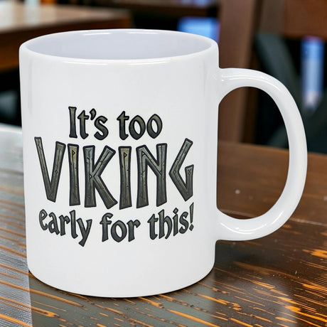 The "It's Too Viking Early For This" 11 oz white ceramic mug by Nordic Wholesale brings Norse humor to your mornings with its bold black lettering, perfect for coffee on a wooden table.