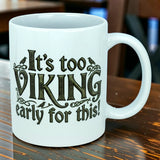 A white 11 oz ceramic mug from Nordic Wholesale sits on a wooden table, showcasing the playful phrase "It's Too Viking Early For This"—a perfect blend of Norse humor to kick off your morning routine.