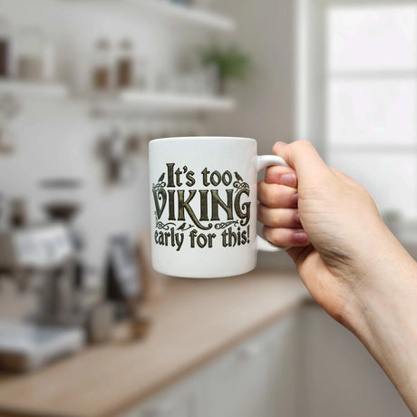 A hand holds the 11 oz "It's Too Viking Early For This" White Ceramic Mug by Nordic Wholesale in a kitchen. The playful Norse-themed quote on the mug stands out, while softly blurred shelves and a window form the background.