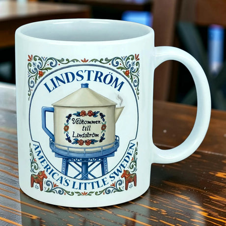 The Nordic Wholesale Mug: Lindstrom America's Little Sweden Ceramic Coffee Mug (11 oz) is a white mug adorned with decorative text that reads “Lindström, America’s Little Sweden” and features an illustration of a water tower bearing the phrase “Välkommen till Lindström,” displayed on a wooden table.
