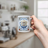 A hand holds a Nordic Wholesale 11 oz ceramic coffee mug featuring a vintage-style water tower design labeled "Lindstrom, America's Little Sweden," with a blurred kitchen background.
