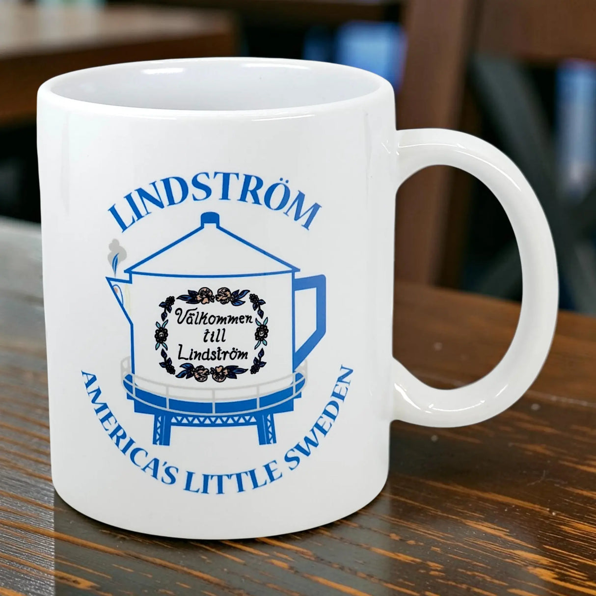The Swedish Gift Store’s 11 oz ceramic mug features a blue teapot design inspired by the Lindstrom Coffee Pot Water Tower, with "America's Little Sweden" and “Välkommen till Lindström” displayed on its white surface.