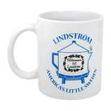 The Swedish Gift Store’s 11 oz ceramic mug features blue "America's Little Sweden" text, the Lindstrom Coffee Pot Water Tower with a “Välkommen till Lindström” sign, and decorative designs—a perfect Chisago Lakes souvenir.