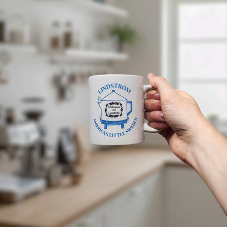 A hand holds a Nordic Wholesale ceramic mug (11 oz) featuring "Lindstrom Coffee Pot Water Tower America's Little Sweden" in blue text and an illustration of the tower, with a blurred kitchen background.