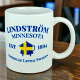 Sure! Here’s a rewritten version using your product data: The Nordic Wholesale Mug: Lindstrom Minnesota America's Little Sweden Ceramic Mug (11 oz Capacity) features blue text reading "Lindström Minnesota EST 1894 America’s Little Sweden" and a Swedish flag shield graphic at the center. Celebrating Lindstrom's rich Swedish heritage, this mug rests on a wooden table.