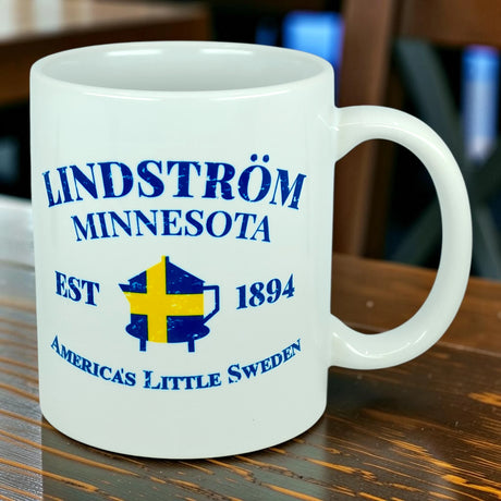 Sure! Here’s a rewritten version using your product data: The Nordic Wholesale Mug: Lindstrom Minnesota America's Little Sweden Ceramic Mug (11 oz Capacity) features blue text reading "Lindström Minnesota EST 1894 America’s Little Sweden" and a Swedish flag shield graphic at the center. Celebrating Lindstrom's rich Swedish heritage, this mug rests on a wooden table.