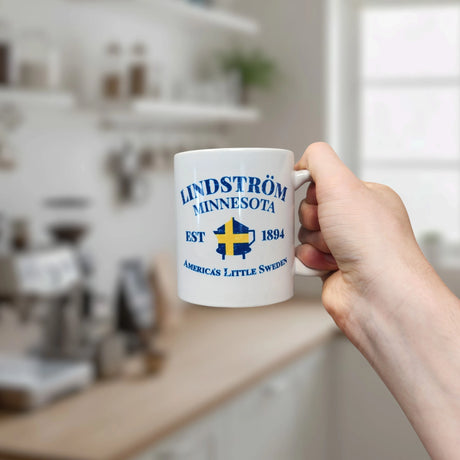 A hand holds a white Nordic Wholesale Mug: Lindstrom Minnesota America's Little Sweden Ceramic Mug (11 oz Capacity) in a kitchen. The mug features the text "Lindström Minnesota Est 1894 America's Little Sweden" with a Swedish flag design beneath it, celebrating its proud Swedish heritage.