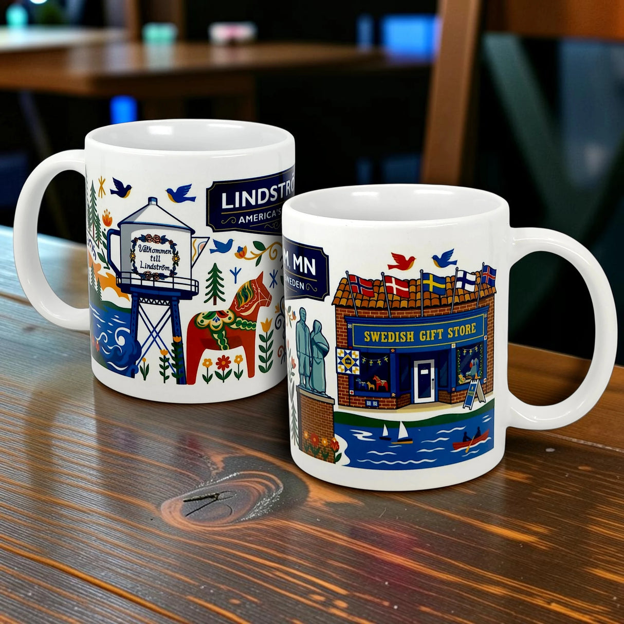 Two 11oz ceramic Lindstrom, MN “America’s Little Sweden” mugs from Swedish Gift Store rest on a wooden table, each featuring colorful illustrations of local landmarks—one with a welcome sign and water tower, the other with people and birds.