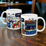 Two 11oz ceramic Lindstrom, MN “America’s Little Sweden” mugs from Swedish Gift Store rest on a wooden table, each featuring colorful illustrations of local landmarks—one with a welcome sign and water tower, the other with people and birds.