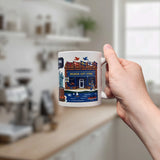 A hand holds the “Mug: Lindstrom, MN — America's Little Sweden” 11oz ceramic souvenir mug by Swedish Gift Store, featuring colorful Swedish scenes, with a bright blurred kitchen in the background.
