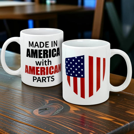 Two white mugs sit on a wooden table: the Nordic Wholesale "Made in America with American Parts" ceramic mug (11 oz), and an American heritage mug with a stylized flag shield—ideal as a Scandinavian-themed gift.