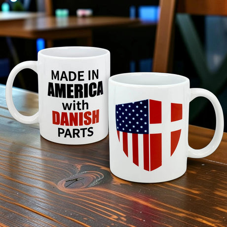 Two white Swedish Gift Store 11 oz ceramic mugs on a wooden table: one says “MADE IN AMERICA with DANISH PARTS” in black and red, the other features a shield combining American and Danish flags.