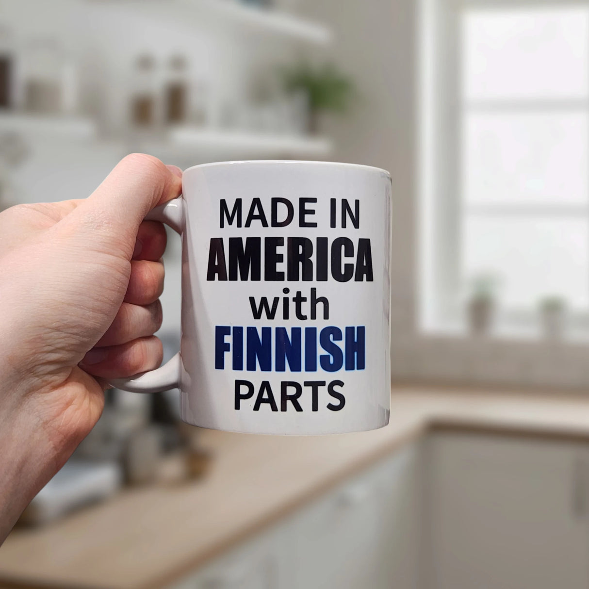 A hand holds the Swedish Gift Store's 11 oz ceramic mug, labeled "Made in America with Finnish Parts," in a modern kitchen with blurred counters and shelves in the background.