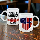 Two white 11 oz ceramic mugs from Swedish Gift Store sit on a wooden table; one reads "Made in America with Norwegian Parts," and the other shows a blended flag shield—a great heritage gift.
