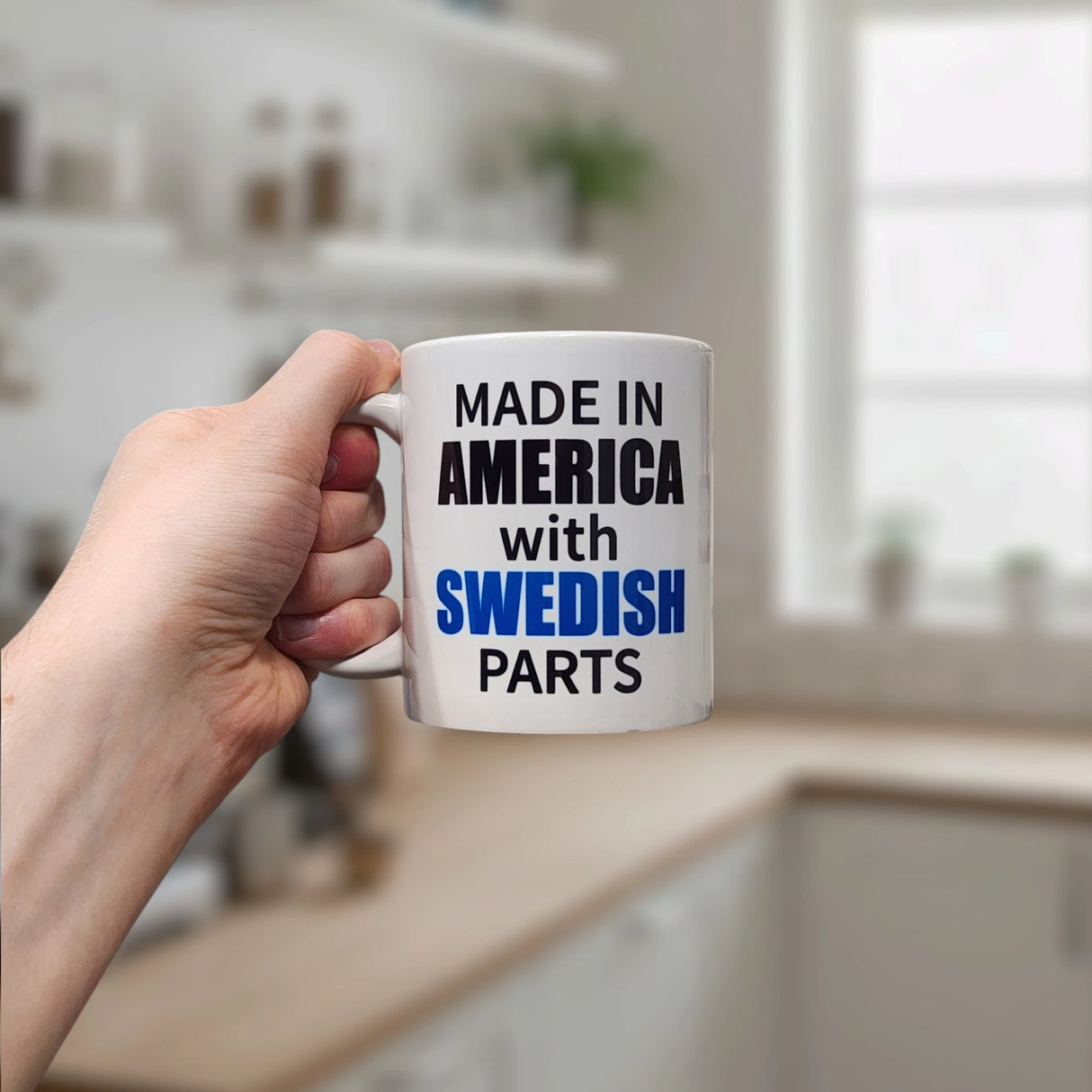 A hand holds the Swedish Gift Store’s 11 oz ceramic mug in a kitchen. The “Made in America with Swedish Parts” design features AMERICA and SWEDISH in bold, while a softly blurred background accentuates the unique look.