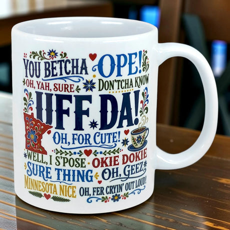 White mug with colorful text in various decorative fonts displaying regional sayings: UFF DA!, YOU BETCHA, OPE!, OKIE DOKIE, and MINNESOTA NICE.