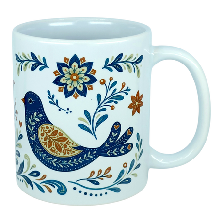 The Nordic Wholesale Mug: Nordic Folk Art Birds Ceramic Mug (11 oz Capacity) is a white Scandinavian-style ceramic mug showcasing a decorative blue bird encircled by detailed floral motifs in blue, orange, and gold hues. It features a plain background and a large handle.