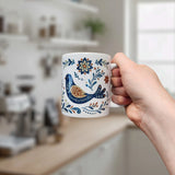 A hand holds the Nordic Wholesale Mug: Nordic Folk Art Birds Ceramic Mug (11 oz Capacity), featuring colorful bird and floral patterns in a white Nordic rosemaling style; a blurred kitchen can be seen in the background.