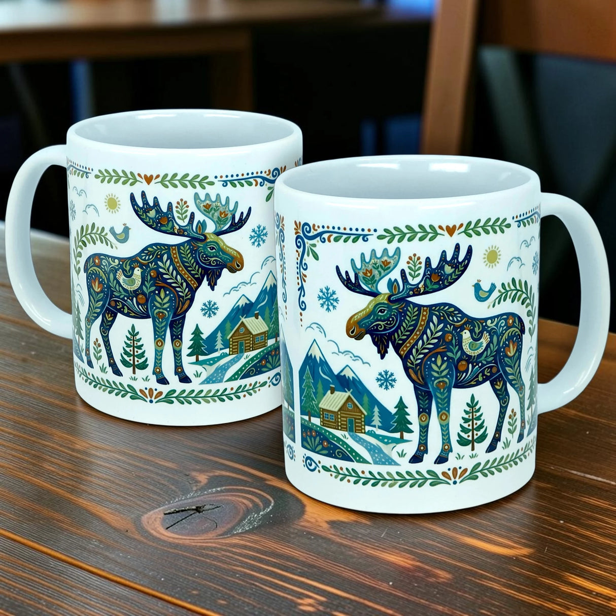 Two Nordic Wholesale ceramic mugs—each a white 11 oz "Nordic Forest Moose" mug—feature vibrant moose designs alongside charming cabin, tree, and mountain motifs, capturing the spirit of Scandinavian folk art as they sit side by side on a wooden table.