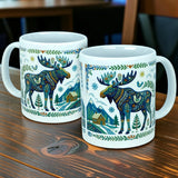 Two Nordic Wholesale ceramic mugs—each a white 11 oz "Nordic Forest Moose" mug—feature vibrant moose designs alongside charming cabin, tree, and mountain motifs, capturing the spirit of Scandinavian folk art as they sit side by side on a wooden table.